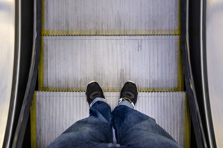 Male feet in jeans and black sneakers standing on escalator POVの写真素材