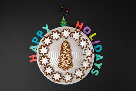 Christmas tree biscuit on white plate with Happy Holidays colorful text on black background - top viewwの写真素材