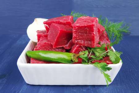slices of raw fresh beef meat fillet in a white bowls with dill and green peppers serving over blue...