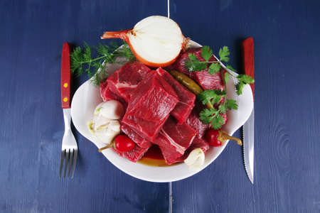 uncooked fresh beef meat chunks on white bowls with vegetables and red peppers serving on blue table...