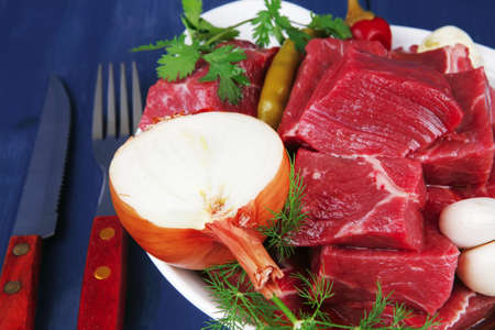 fresh uncooked beef meat slices over white bowls ready to prepare with red peppers and greenery serving on blue table with cutleryの写真素材