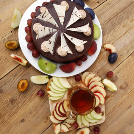 big whole frost chocolate cream brownie cake topped with white chocolate and cream flowers with hot tea cup decorated with fruits apple plum and grape on plate on wooden tableの写真素材