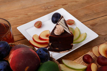 slice of chocolate cream brownie cake topped with white chocolate and cream flowers with hot tea mug decorated with fruits apple plum and grape on plate on wooden tableの写真素材