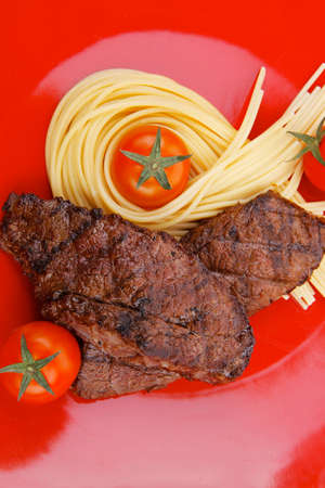 meat grilled beef steak with pasta and tomatoes on red plate isolated over white backgroundの写真素材