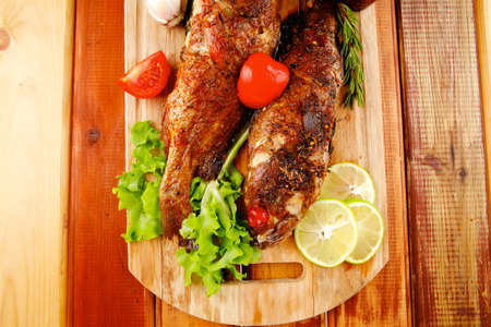 healthy food: two fried sea bass fish served with tomatoes and vegetables on big wooden board over...