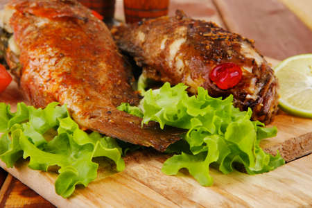 healthy food: two fried sea bass fish served with tomatoes and vegetables on big wooden board over...