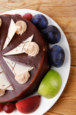 chocolate cream brownie cake topped with white chocolate slice and cream flowers decorated with fruits apple plum and grape on plate on wooden tableの写真素材