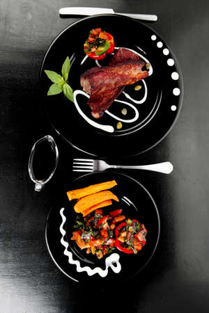 fresh red beef meat steak barbecue garnished vegetable salad sweet potato and basil on black plate over black wooden table with bbq sauce in sauceboatの写真素材