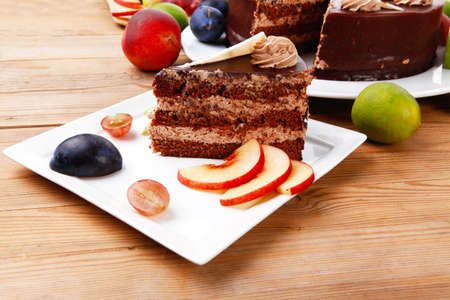chocolate cream brownie cake layers slice topped with white chocolate and cream flowers decorated with fruits apple plum and grape on plate on wooden table with big whole cakeの写真素材