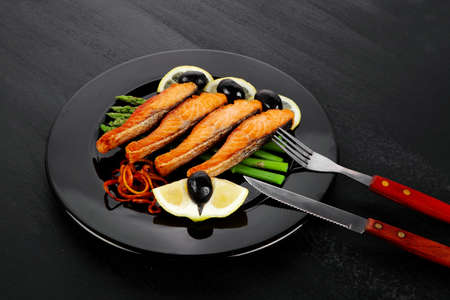 grilled salmon slices with asparagus lemon olives and cutlery on black plate over dark wooden tableの写真素材