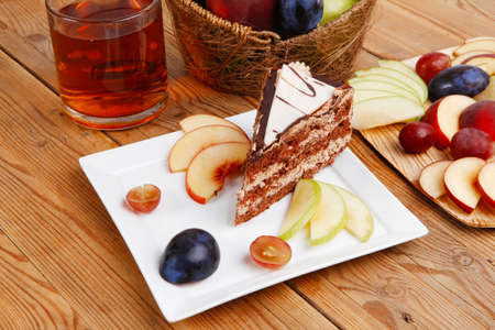 slice of chocolate cream brownie cake topped with white chocolate and cream flowers with hot tea mug decorated with fruits apple plum and grape on plate on wooden tableの写真素材