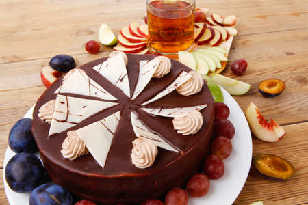 big whole frost chocolate cream brownie cake topped with white chocolate and cream flowers with hot tea cup decorated with fruits apple plum and grape on plate on wooden tableの写真素材