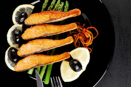 grilled salmon slices with asparagus lemon olives and cutlery on black plate over dark wooden table