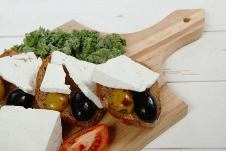 fresh greek feta cheese sandwich on wooden plate with hot green olives and black, goat cube, tomatoes kale over tableの写真素材