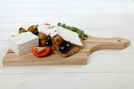fresh greek feta cheese sandwich on wooden plate with hot green olives and black, goat cube, tomatoes kale over tableの写真素材