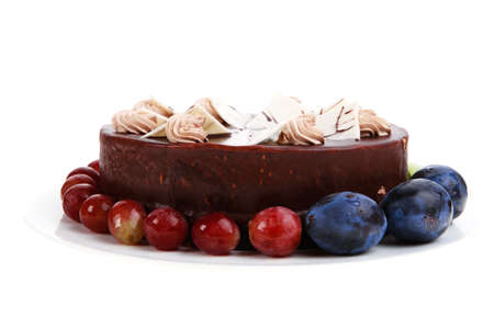 chocolate cream brownie cake topped with white chocolate slice and cream flowers decorated with fruits apple plum and grape on plate isolated over white backgroundの写真素材