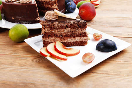 chocolate cream brownie cake layers slice topped with white chocolate and cream flowers decorated with fruits apple plum and grape on plate on wooden table with big whole cakeの写真素材