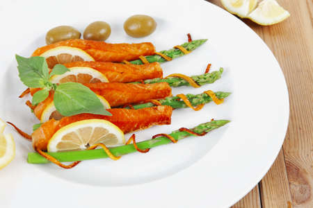 roasted salmon slices with asparagus lemon fried orange peel green olives and cutlery on white plate over wooden tableの写真素材