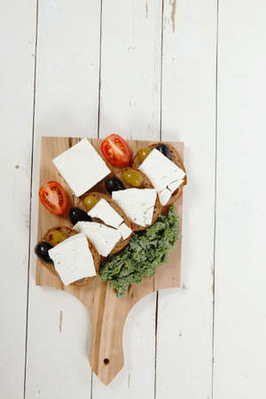 fresh greek feta cheese sandwich on wooden plate with hot green olives and black, goat cube, tomatoes kale over tableの写真素材