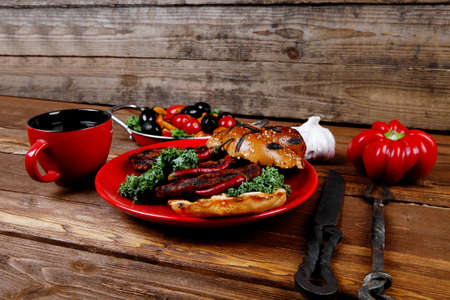 fresh grilled beef hamburger served on red plate with black coffee glass chili pepper olives green kale leaves mushroom bell garlic and forged vintage antique cutlery over wooden tableの写真素材