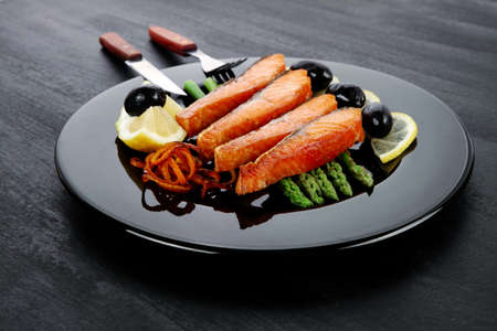 grilled salmon slices with asparagus lemon olives and cutlery on black plate over dark wooden tableの写真素材