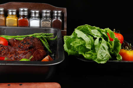 fresh glazed baked big beef meat rib chunk under sweet sauce with lettuce broccoli tomatoes hot chili pepper pink peppercorn different spices on black wooden tableの写真素材