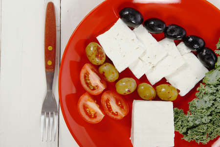 fresh greek feta cheese sandwich on red plate with hot green olives and black, goat cheese cube, tomatoes kale over retro tableの写真素材