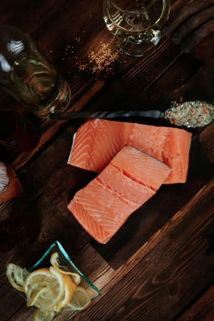 fresh raw salmon fillet served with lemon and white wine in wineglass, bottle, on dark plate over vintage wood table with handmade cutlery knife and spice on spoonの写真素材