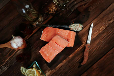 fresh raw salmon fillet served with lemon and white wine in wineglass, bottle, on dark plate over vintage wood table with handmade cutlery knife and spice on spoonの写真素材