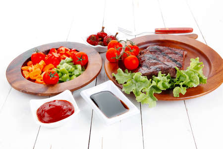 dinner of fresh rich juicy grilled beef meat steak fillet with marks on wooden plate over white table served with vegetable salad and cutlery, new york styled cuisineの写真素材