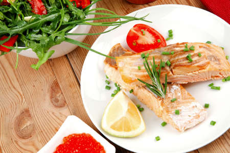 savory fish portion : roasted norwegian salmon chunks with  lemon and vegetable salad , rosemary twig and red caviar in white bowl over wooden tableの写真素材
