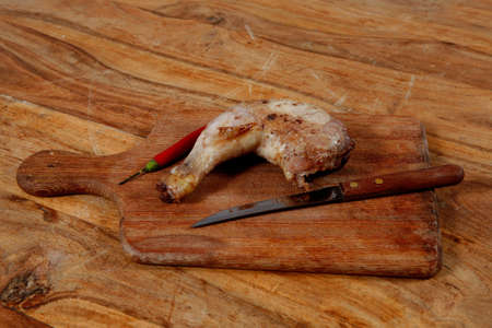 grilled chicken thighs on wooden cut board and red hot chili pepper and stainless steel knife over light wooden tableの写真素材