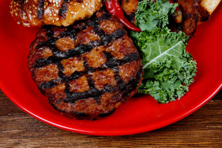 fresh grilled beef hamburger served on red plate with black coffee glass chili pepper olives green kale leaves mushroom bell garlic and forged vintage antique cutlery over wooden tableの写真素材