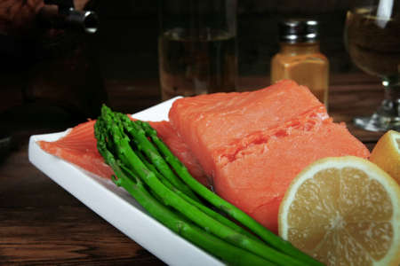 fresh raw salmon fillet served with lemon and white wine in wineglass, wooden barrel, bottle, on white plate over vintage wood table with handmade cutlery knife and forkの写真素材