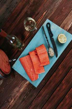 fresh raw salmon fillet served with lemon and white wine in wineglass bottle wooden barrel on blue plate over vintage wood table with forged handmade cutlery knife and spice on spoonの写真素材