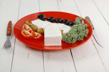 fresh greek feta cheese sandwich on red plate with hot green olives and black, goat cheese cube, tomatoes kale over retro tableの写真素材