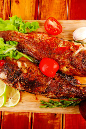 healthy food: two fried sea bass fish served with tomatoes and vegetables on big wooden board over...