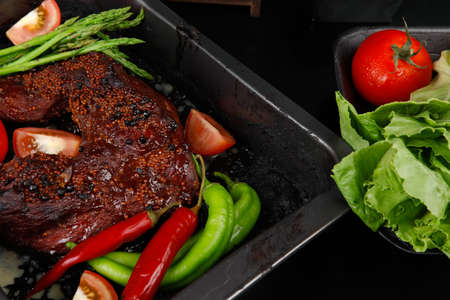 fresh glazed baked big beef meat rib chunk under sweet sauce with lettuce broccoli tomatoes hot chili pepper pink peppercorn different spices on black wooden tableの写真素材