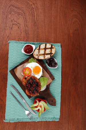 junk food big beef hamburger fried eggs on dark wood plate with modern cutlery ketchup sauce and pickels on blue mat isolated over white backgroundの写真素材