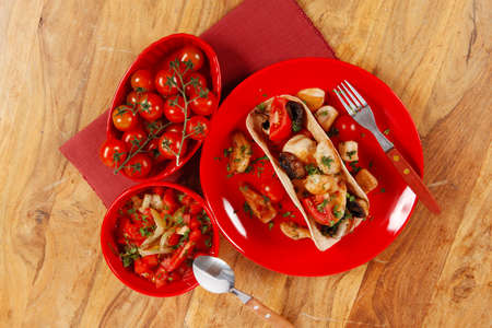 big mexican taco with tomatoes and mushroom, served with soup, fresh vegetables , on red crockery plates over wooden table with cutlery and napkinの写真素材