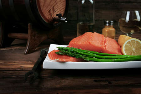 fresh raw salmon fillet served with lemon and white wine in wineglass, wooden barrel, bottle, on white plate over vintage wood table with handmade cutlery knife and forkの写真素材
