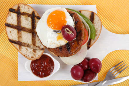junk food meat big beef hamburger fried eggs on white wood plate with cutlery ketchup sauce and pickels on yellow mat isolated over white backgroundの写真素材