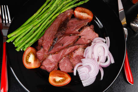 business lunch fresh roast beef meat slices on black plate wotj cutlery asparagus boiled broccoli rye bun on wooden tableの写真素材