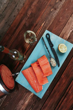 fresh raw salmon fillet served with lemon and white wine in wineglass bottle wooden barrel on blue plate over vintage wood table with forged handmade cutlery knife and 
spice on spoonの写真素材