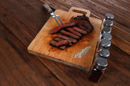grilled beef fillet steak on fork over wooden board served with different kind of dry spices salt crushed black pepper hot baked sweet paprika red hot chili on dark tableの写真素材