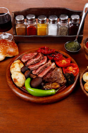fresh roast bbq beef meat ribeye steak on wooden plate served with tomato juice in wooden cup, boiled broccoli, baked tomatoes and potatoes, with white bun, and red wine glass on light walnut wooden tableの写真素材