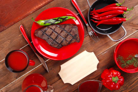red theme lunch : fresh grilled bbq roast beef steak on red plate with green chili tomato soup ketchup sauce paprika small jug glass ground pepper american peppercorn and modern cutlery served on wooden table with empty nameplateの写真素材