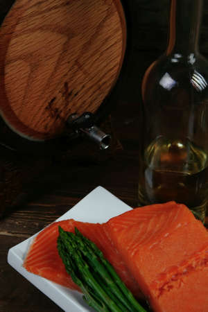fresh raw salmon fillet served with lemon and white wine in wineglass, wooden barrel, bottle, on white plate over vintage wood table with handmade cutlery knife and forkの写真素材