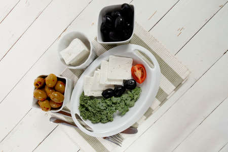 fresh greek feta cheese sandwich on white plate with hot green olives and black, goat cheese cube, tomatoes kale over retro tableの写真素材