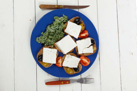 white feta greek cheese sandwich rye bread on blue plate with black spain olives tomatoes and cutlery over retro wooden tableの写真素材